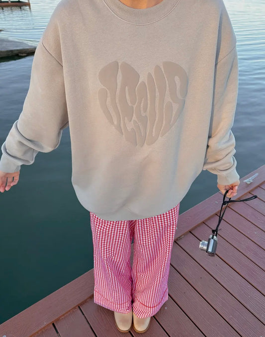 Person wearing a beige sweatshirt with a Jesus heart design and pink checkered pants on a dock.