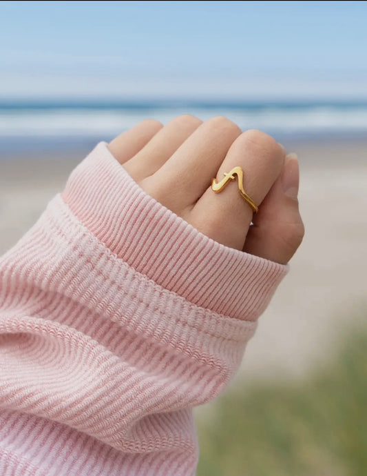 Hand wearing a gold Wavy Cross ring with a pink sleeve against a blurred natural background