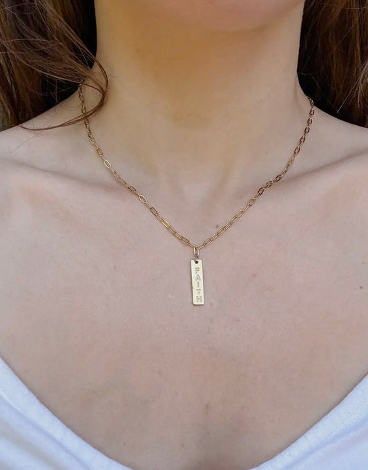 Gold necklace with a bar pendant with the word faith on a person wearing a white top.