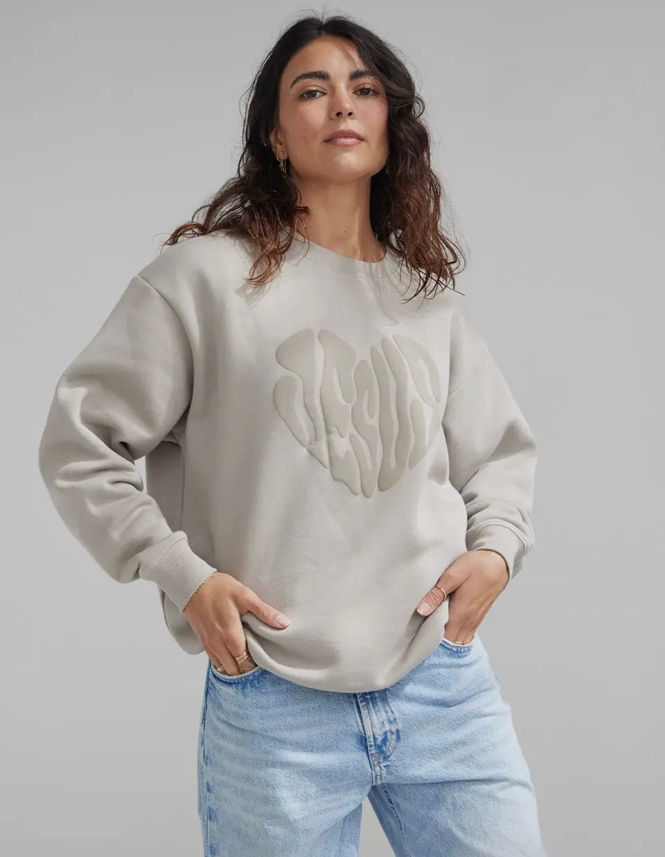 Woman wearing a beige sweatshirt with a Jesus heart logo and light blue jeans on a gray background
