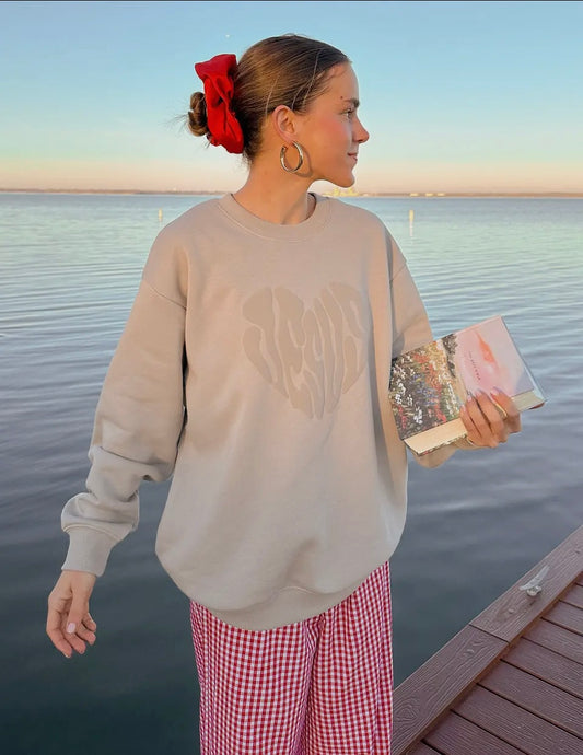 Woman standing by a waterfront wearing a beige sweatshirt and red checkered pants, holding a clutch.