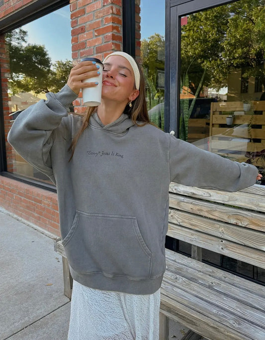 Woman wearing a gray hoodie with text, “sorry, Jesus is King” standing outdoors near a building.