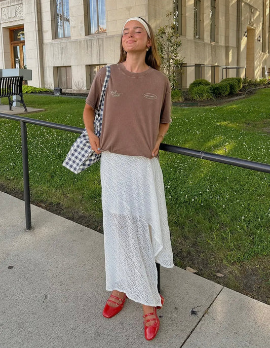 Woman in a brown t-shirt and white lace skirt standing on a sidewalk with a building in the background.