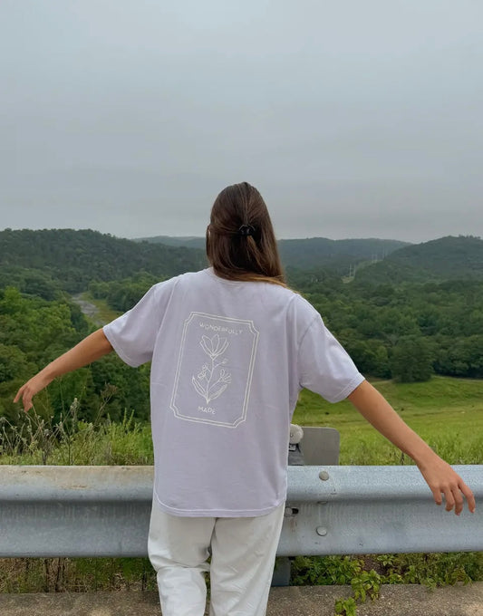 Person wearing a white t-shirt with a design on the back, standing with arms outstretched over a scenic view of green hills.