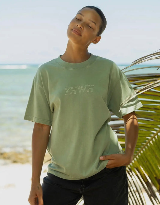 Person wearing a light green t-shirt with 'YHWH' printed on it, standing by a beach.