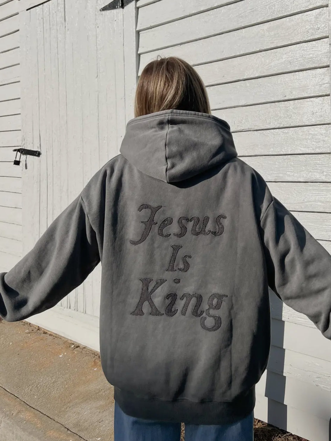 Person wearing a hoodie with 'Jesus Is King' text on the back, standing in front of a wooden door.